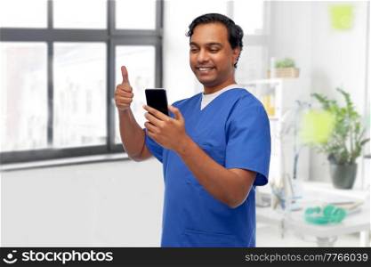medicine, healthcare and technology concept - happy smiling doctor or male nurse in blue uniform having video call on smartphone and showing thumbs up over medical office at hospital background. doctor or male nurse having video call on phone