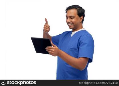 medicine, healthcare and technology concept - happy smiling indian doctor or male nurse in blue uniform having video call on tablet pc computer and showing thumbs up over white background. doctor or male nurse has video call on tablet pc