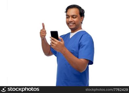 medicine, healthcare and technology concept - happy smiling indian doctor or male nurse in blue uniform having video call on smartphone and showing thumbs up over white background. doctor or male nurse having video call on phone