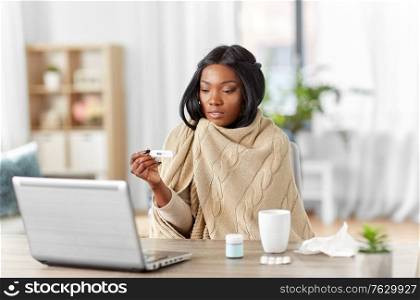 medicine, healthcare and technology concept - sick young african american woman having video call or online consultation on laptop computer and measuring temperature with thermometer at home. sick woman having video call on laptop at home
