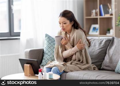 medicine, healthcare and technology concept - sick young asian woman having video call or online consultation on tablet pc computer and drinking tea at home. sick woman having video call on tablet pc at home