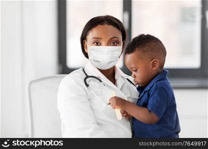 medicine, healthcare, pediatry and people concept - african american female doctor or pediatrician wearing face protective mask holding baby boy patient on medical exam at clinic. doctor in mask with baby patient at clinic