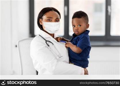 medicine, healthcare, pediatry and people concept - african american female doctor or pediatrician wearing protective mask holding baby boy patient on medical exam at clinic. doctor in mask with baby patient at clinic