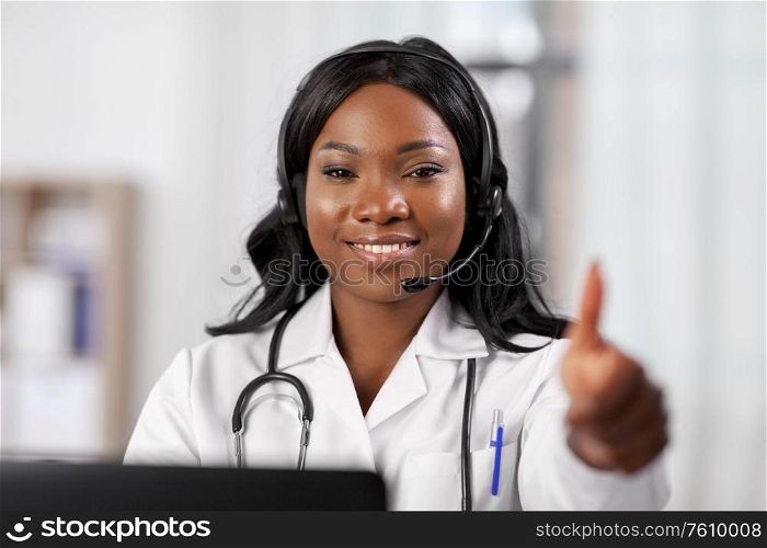 medicine, online service and healthcare concept - happy smiling african american female doctor or nurse with headset and laptop at hospital showing thumbs up. african doctor with headset and laptop at hospital