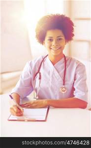medicine, people and healthcare concept - happy female african american doctor or nurse writing medical report to clipboard at hospital