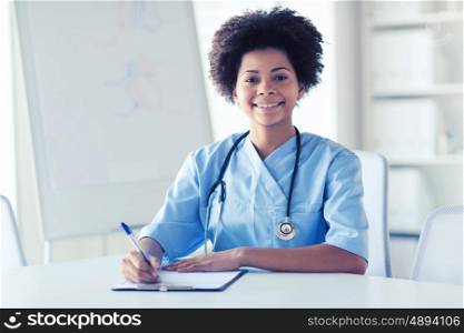 medicine, people and healthcare concept - happy female african american doctor or nurse writing medical report to clipboard at hospital