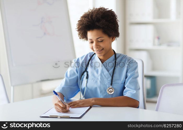 medicine, people and healthcare concept - happy female african american doctor or nurse writing medical report to clipboard at hospital