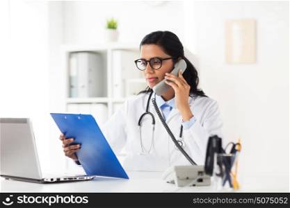 medicine, people and healthcare concept - happy female doctor with clipboard and laptop computer calling on phone at hospital. doctor with clipboard calling on phone at hospital