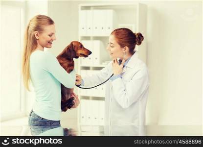 medicine, pet, animal, health care and people concept - happy woman holding dachshund and veterinarian doctor with stethoscope checking dog heartbeat at vet clinic