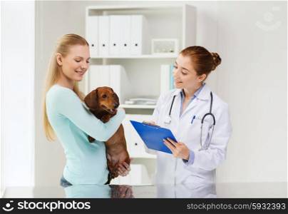 medicine, pet care and people concept - happy woman holding dachshund dog and veterinarian doctor with clipboard at vet clinic