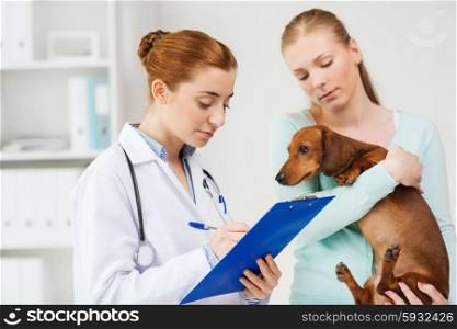 medicine, pet care and people concept - woman holding dachshund dog and veterinarian doctor with clipboard taking notes at vet clinic