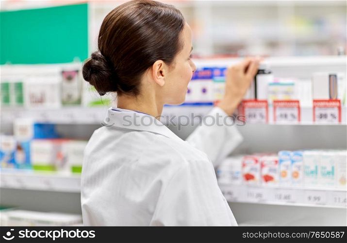 medicine, pharmaceutics, health care and people concept - female pharmacist at shelf with drugs at pharmacy. female pharmacist with medicine at pharmacy