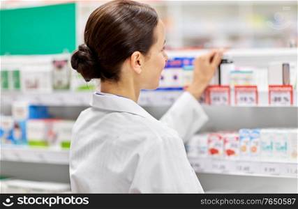 medicine, pharmaceutics, health care and people concept - female pharmacist at shelf with drugs at pharmacy. female pharmacist with medicine at pharmacy