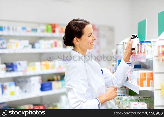 medicine, pharmaceutics, healthcare and people concept - happy female apothecary taking drug from shelf at pharmacy. happy female apothecary with drug at pharmacy