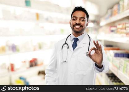 medicine, pharmacy and healthcare concept - smiling indian male doctor or pharmacist in white coat with stethoscope showing ok hand sign over drugstore background. smiling indian male doctor showing ok gesture
