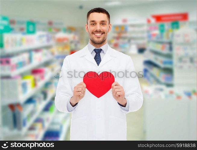 medicine, pharmacy, people, health care and pharmacology concept - happy male pharmacist holding red heart shape over drugstore background