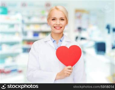 medicine, pharmacy, people, health care and pharmacology concept - happy young woman pharmacist holding red heart shape over drugstore background