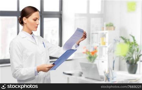 medicine, profession and healthcare concept - female doctor in white coat with clipboard reading medical report over medical office at hospital on background. female doctor with medical report at hospital