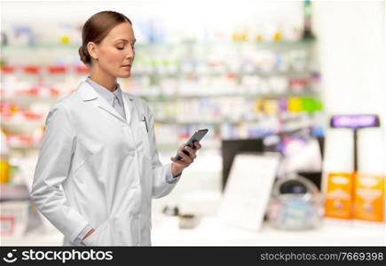 medicine, profession and healthcare concept - female doctor or pharmacist with stethoscope using smartphone over pharmacy background. female doctor with smartphone at pharmacy
