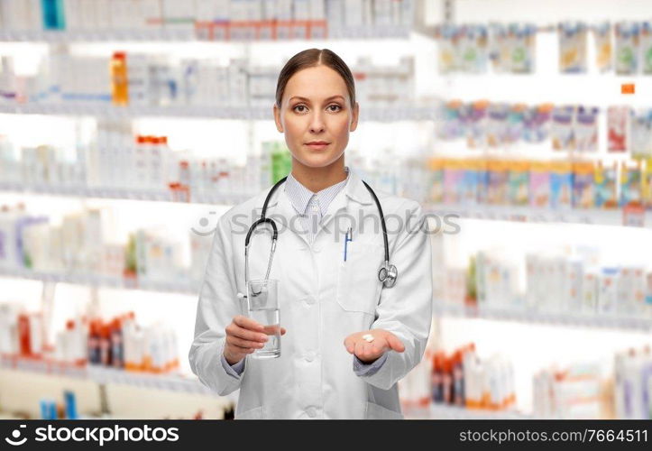 medicine, profession and healthcare concept - female doctor with pill and glass of water over pharmacy background. doctor with pills and glass of water at pharmacy