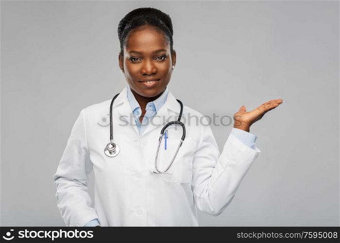medicine, profession and healthcare concept - happy smiling african american female doctor in white coat with stethoscope showing something imnaginary over background. african american female doctor with stethoscope