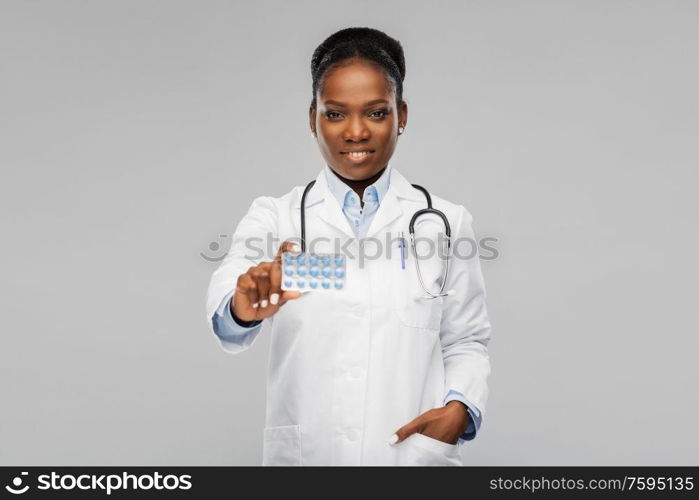 medicine, profession and healthcare concept - happy smiling african american female doctor or in white coat with pills and stethoscope over background. african american female doctor with medicine pills