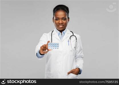 medicine, profession and healthcare concept - happy smiling african american female doctor or in white coat with pills and stethoscope over background. african american female doctor with medicine pills