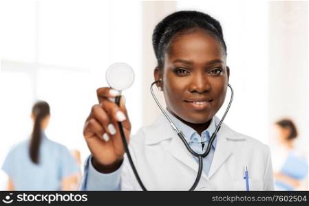 medicine, profession and healthcare concept - happy smiling african american female doctor in white coat with stethoscope over hospital staff on background. african american female doctor with stethoscope