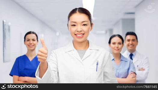 medicine, profession and healthcare concept - happy smiling asian female doctor in white coat showing thumbs up with medical team at hospital on background. asian female doctor showing thumbs up at hospital