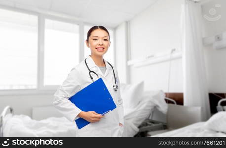 medicine, profession and healthcare concept - happy smiling asian female doctor in white coat with clipboard and pen over hospital background. asian female doctor with clipboard at hospital