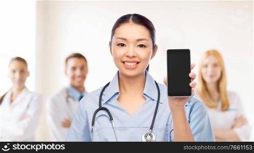 medicine, profession and healthcare concept - happy smiling asian female doctor or nurse in blue uniform with stethoscope showing smartphone with medical team at hospital on background. happy asian female doctor or nurse with smartphone