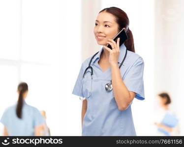 medicine, profession and healthcare concept - happy smiling asian female doctor or nurse in blue uniform with stethoscope calling on smartphone over hospital background. asian nurse calling on smartphone at hospital