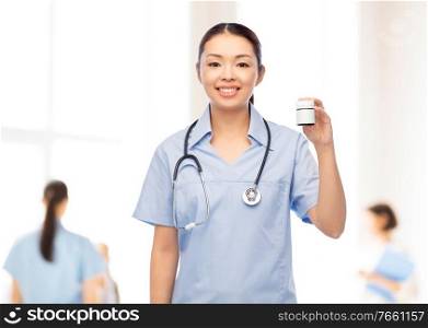 medicine, profession and healthcare concept - happy smiling asian female doctor or nurse in blue uniform with stethoscope holding jar of pills over hospital background. happy asian female nurse with medicine at hospital