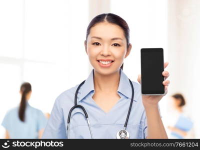 medicine, profession and healthcare concept - happy smiling asian female doctor or nurse in blue uniform with stethoscope showing smartphone over white background. asian female nurse with smartphone at hospital