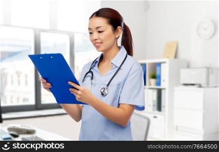 medicine, profession and healthcare concept - happy smiling asian female doctor or nurse in blue uniform with clipboard and pen over hospital background. asian female doctor with clipboard at hospital
