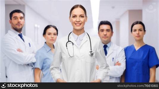 medicine, profession and healthcare concept - happy smiling female doctor in white coat with stethoscope over group of colleagues at hospital on background. smiling female doctor with colleagues at hospital