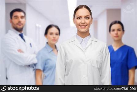 medicine, profession and healthcare concept - happy smiling female doctor in white coat over group of colleagues at hospital on background. smiling female doctor with colleagues at hospital