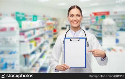 medicine, profession and healthcare concept - happy smiling female doctor in white coat showing clipboard over pharmacy background. happy female doctor with clipboard at pharmacy