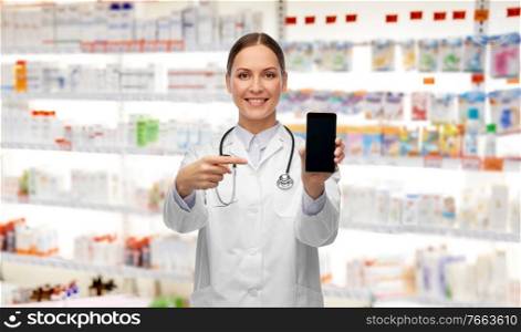 medicine, profession and healthcare concept - happy smiling female doctor or nurse with stethoscope showing smartphone over pharmacy background. happy female doctor or nurse with smartphone