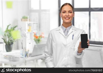 medicine, profession and healthcare concept - happy smiling female doctor showing smartphone over white background over medical office at hospital on background. smiling female doctor with smartphone at hospital