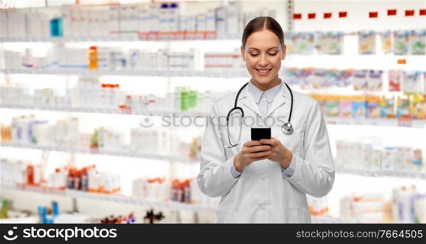 medicine, profession and healthcare concept - happy smiling female doctor with stethoscope using smartphone over pharmacy background. happy smiling female doctor with smartphone