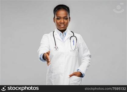 medicine, profession and healthcare concept - smiling african american female doctor in white coat and stethoscope giving hand for handshake over grey background. african american female doctor making handshake