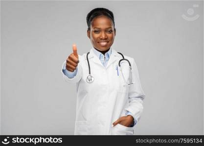 medicine, profession and healthcare concept - smiling african american female doctor in white coat with stethoscope showing thumbs up over grey background. african american female doctor showing thumbs up
