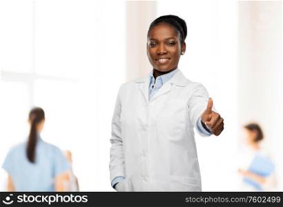 medicine, profession and healthcare concept - smiling african american female doctor or scientist in white coat showing thumbs up over hospital staff on background. african american female doctor showing thumbs up