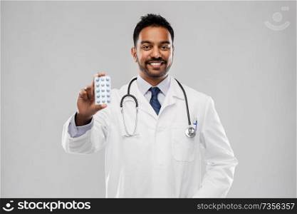 medicine, profession and healthcare concept - smiling indian male doctor in white coat with pills and stethoscope over grey background. indian male doctor with pills and stethoscope