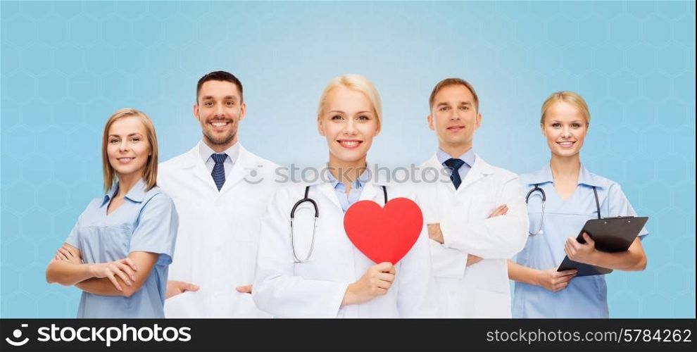 medicine, profession, teamwork and healthcare concept - group of smiling medics or doctors holding red paper heart shape, clipboard and stethoscopes over blue background