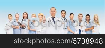 medicine, profession, teamwork and healthcare concept - international group of smiling medics or doctors with clipboard and stethoscopes with showing thumbs up over blue background