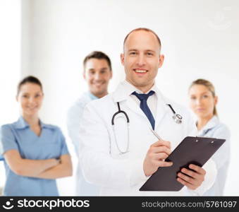 medicine, profession, teamwork and healthcare concept - smiling male doctor with clipboard and stethoscope writing prescription over group of medics