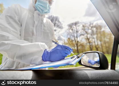 medicine, quarantine and pandemic concept - close up of doctor or healthcare worker in protective gear or hazmat suit, medical mask, face shield and goggles writing to clipboard at car. doctor in hazmat suit with clipboard at car
