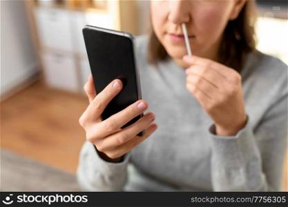 medicine, quarantine and pandemic concept - close up of woman with swab and smartphone taking s&le from her nose and making nasal coronavirus self test at home. woman making self testing coronavirus test at home
